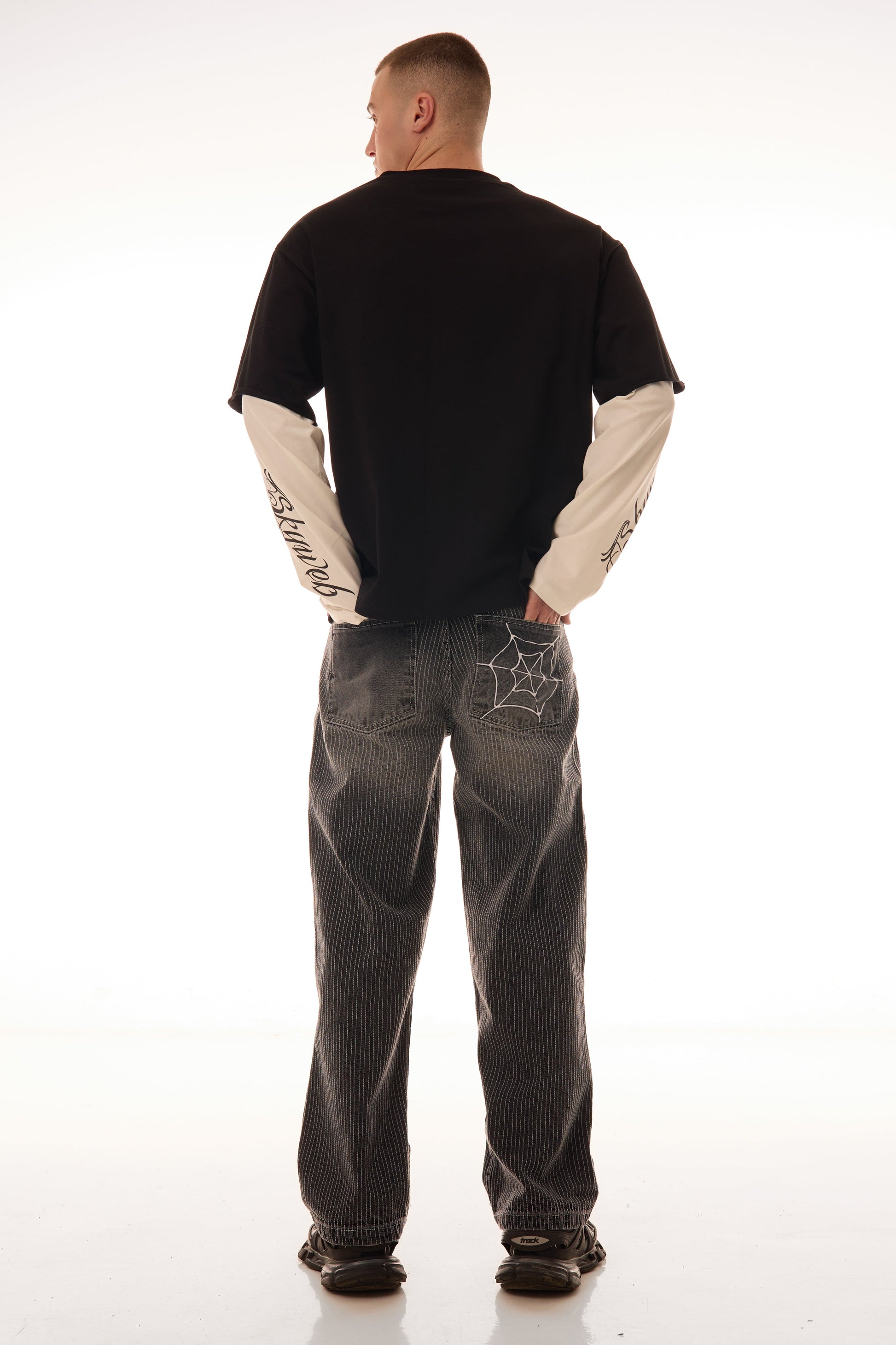 Model văzut din spate purtând blugii Axis Denim gri, wide fit, cu tighele verticale albe și broderie pe buzunarul spate.
