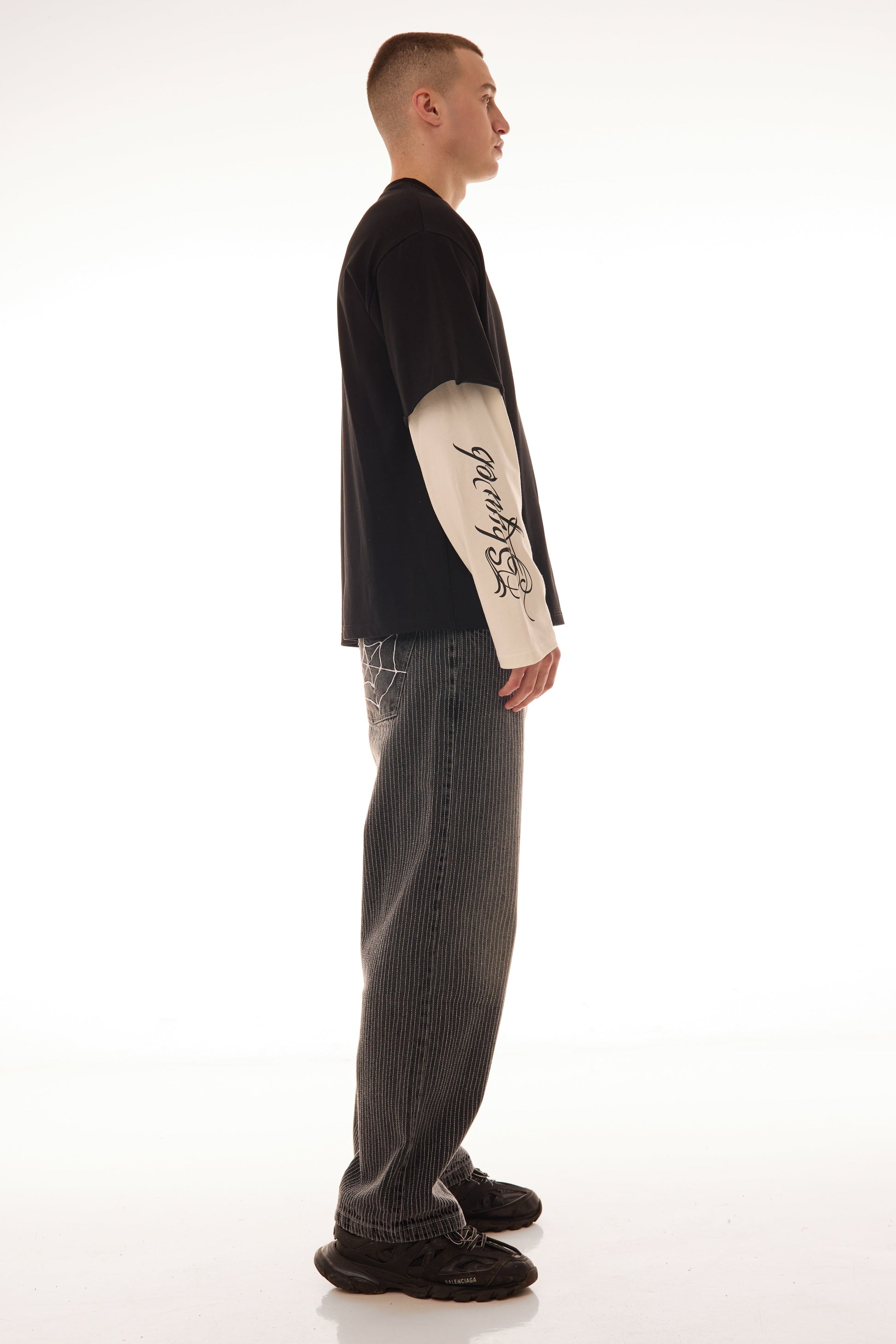 Model văzut din lateral purtând tricoul DUAL LONG TEE Skyweb, tricou negru cu mâneci albe layered și print gotic pe mâneci.