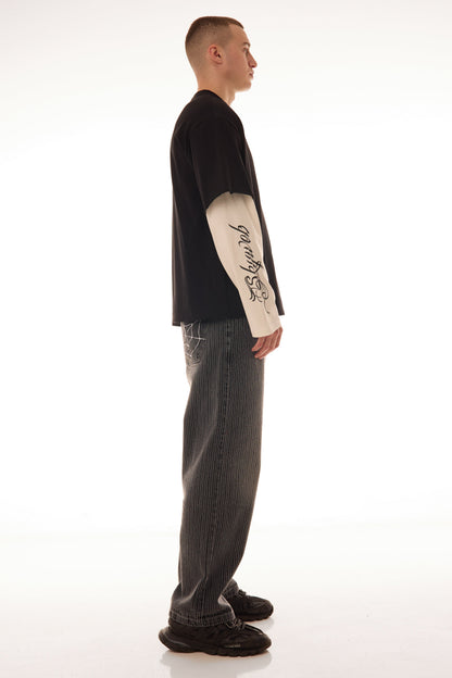 Model văzut din lateral purtând tricoul DUAL LONG TEE Skyweb, tricou negru cu mâneci albe layered și print gotic pe mâneci.