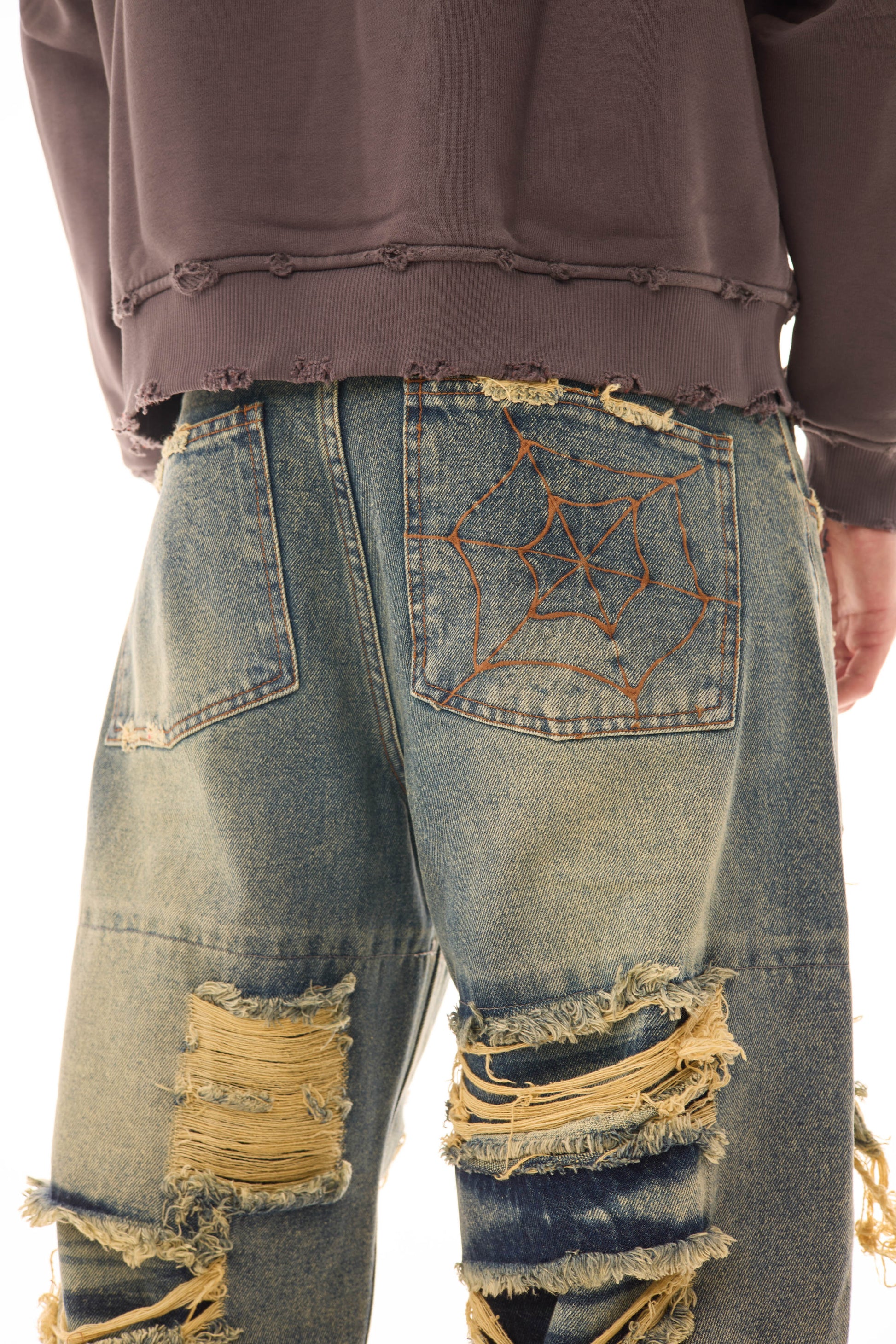 Detaliu spate al blugilor Shattered Denim Blue 13.5 OZ, wide fit, cu rupturi mari și broderie tip pânză de păianjen pe buzunarul din spate.
